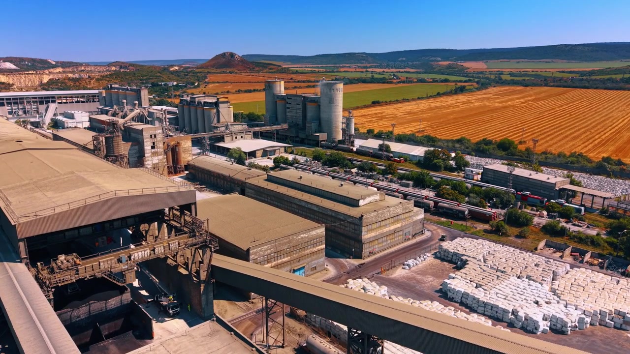 Fabbrica industriale accanto a campi mietuti. Grandi edifici di produzione e nastri trasportatori chiusi si trovano accanto a silos, vagoni ferroviari, pacchi immagazzinati e terreni agricoli.