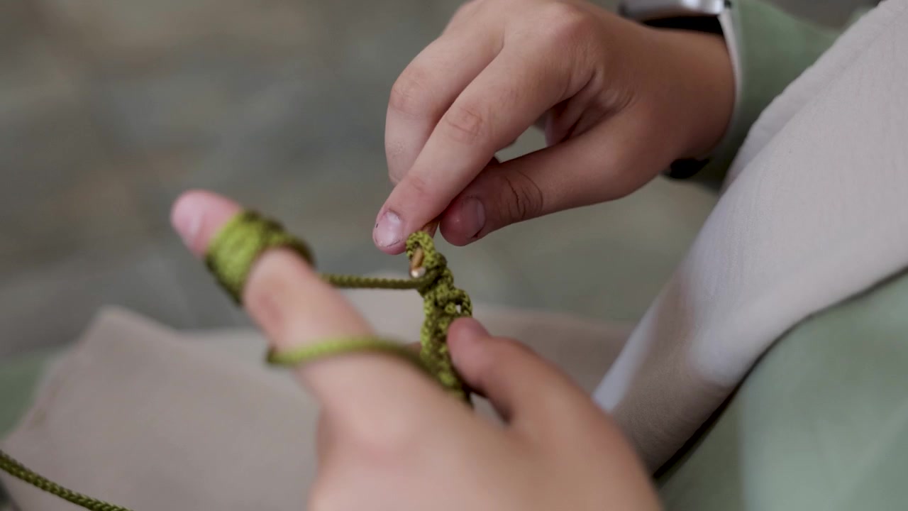 Close-up of a woman’s hands crocheting green yarn with a hook, showing the detailed handmade process of textile craftsmanship.