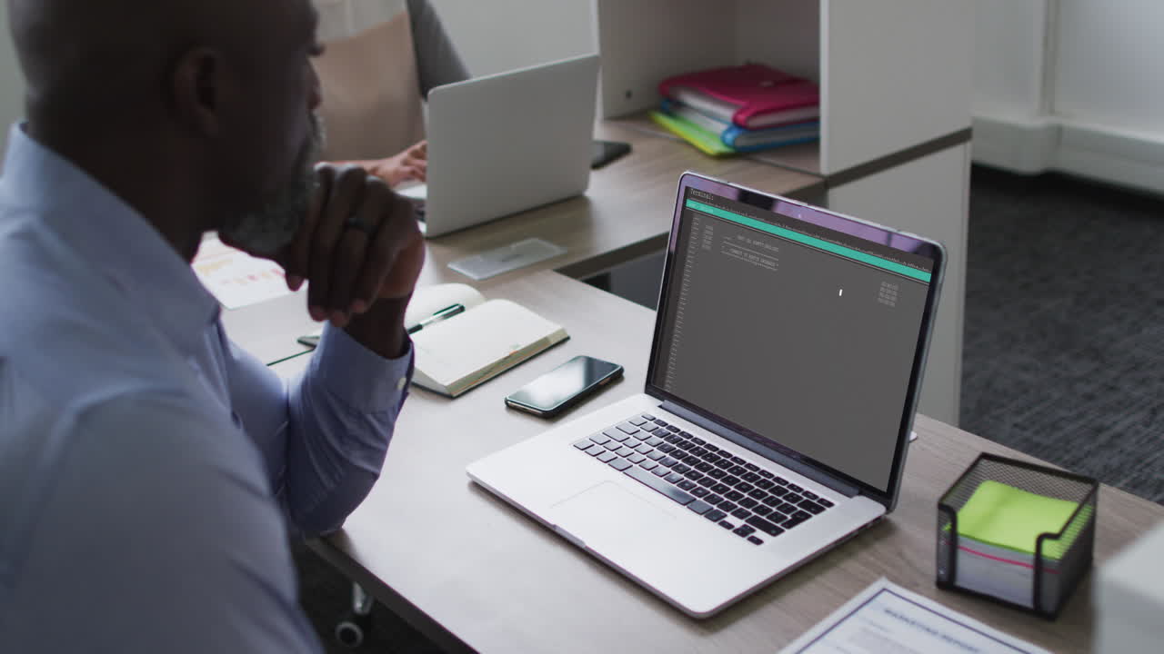 African american man sitting at desk watching coding data processing on laptop screen