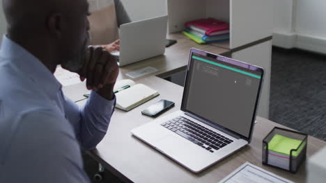hombre afroamericano sentado en el escritorio viendo el procesamiento de datos de codificación en la pantalla del portátil