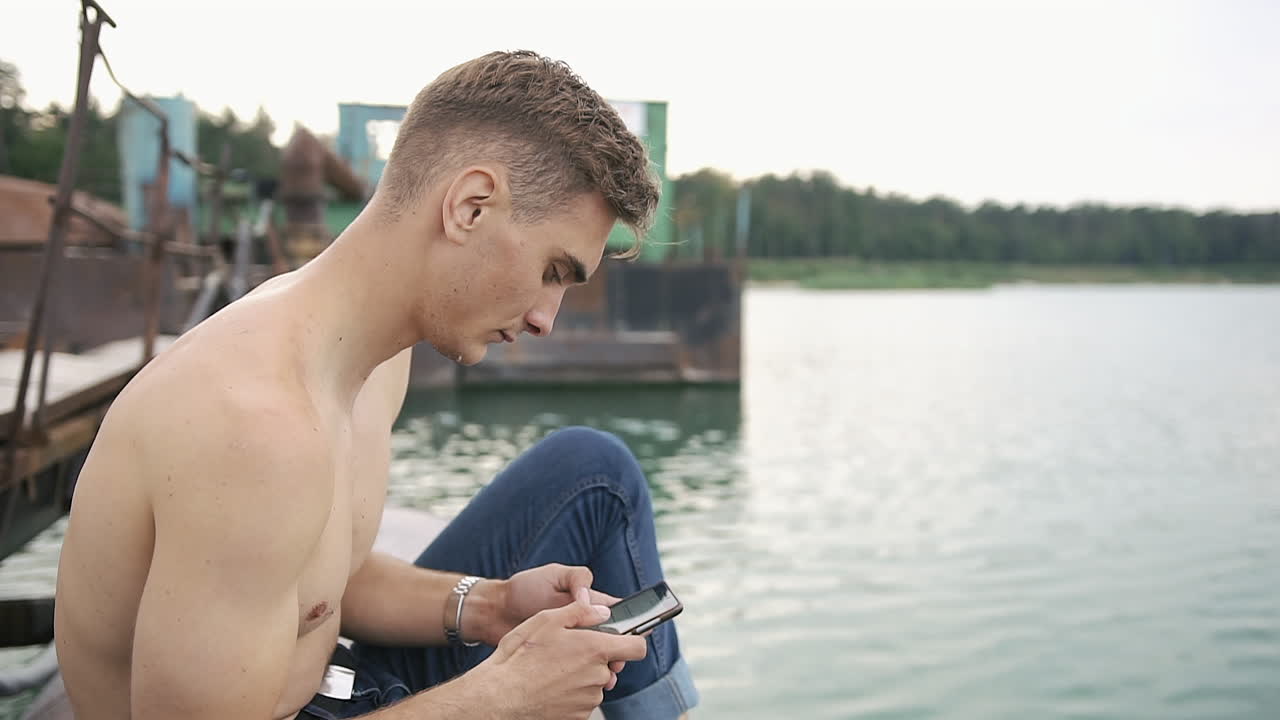 Shirtless man using smartphone by the lake