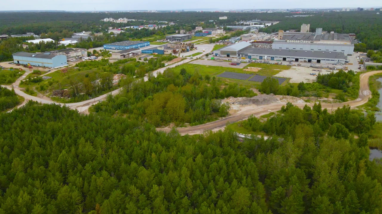 Aerial drone footage flying above large industrial warehouses and workshop buildings in a Männiku forest during a cloudy day. Treetops and gravel roads around the facility. Storage containers visible.