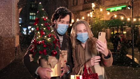 caucasian happy couple wearing facial masks holding christmas tree and laughing while making a selfie with smartphone on the street in christmas