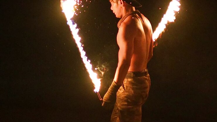 A shirtless performer skillfully spins a flaming staff in the dark, showcasing dynamic movement and fiery sparks.