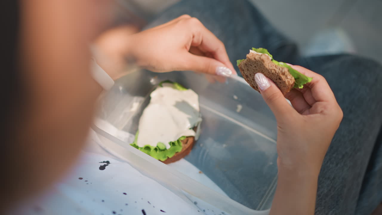 Una mujer preparando un bocado en un recipiente, una persona que viaja al trabajo cogiendo un almuerzo portátil en el regazo con el reloj a la vista, untado cremoso y lechuga en pan con semillas, ambiente urbano dinámico y primer plano con poca profundidad, los fotogramas retratan