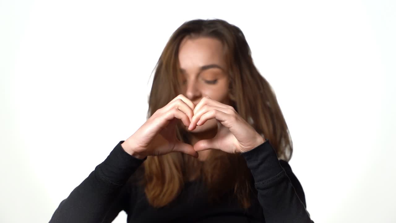 Woman is portraying heart by her hands before her face on a white background in the studio. Close-up