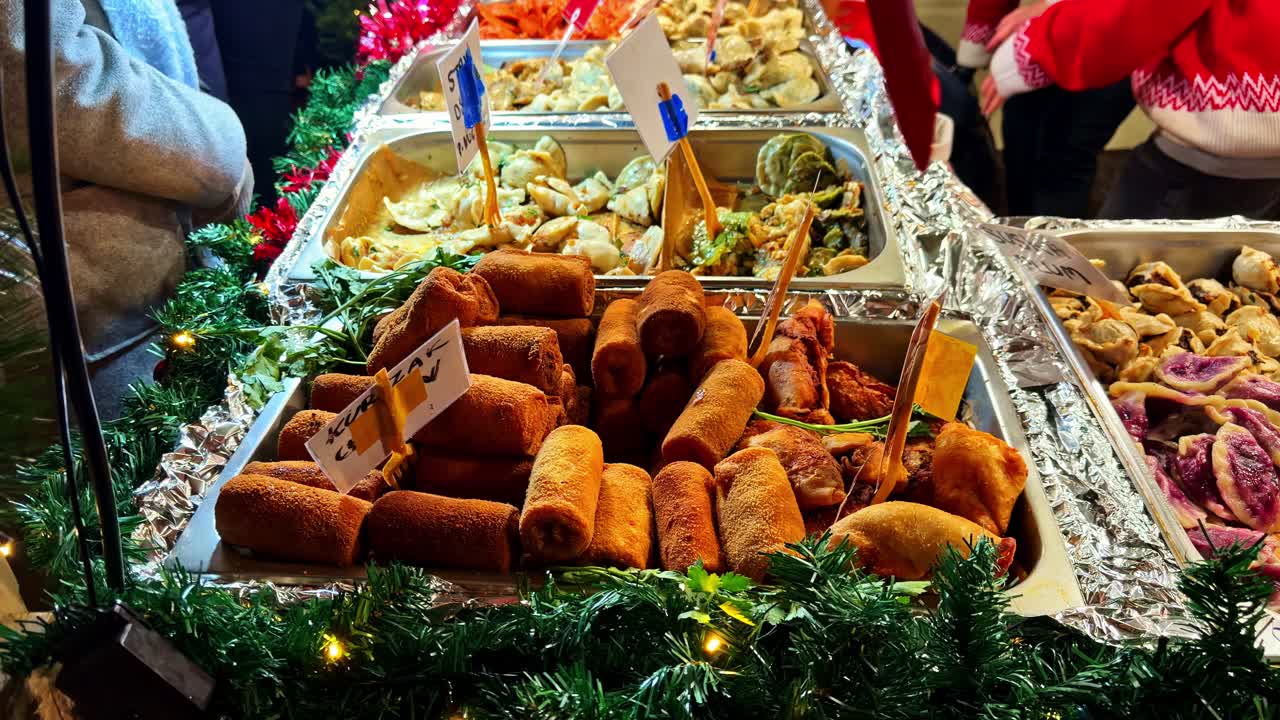 Primo piano di vassoi con crocchette impanate e ravioli pierogi in una bancarella di street food festiva, incorniciati da ghirlande natalizie e luci calde per i visitatori invernali