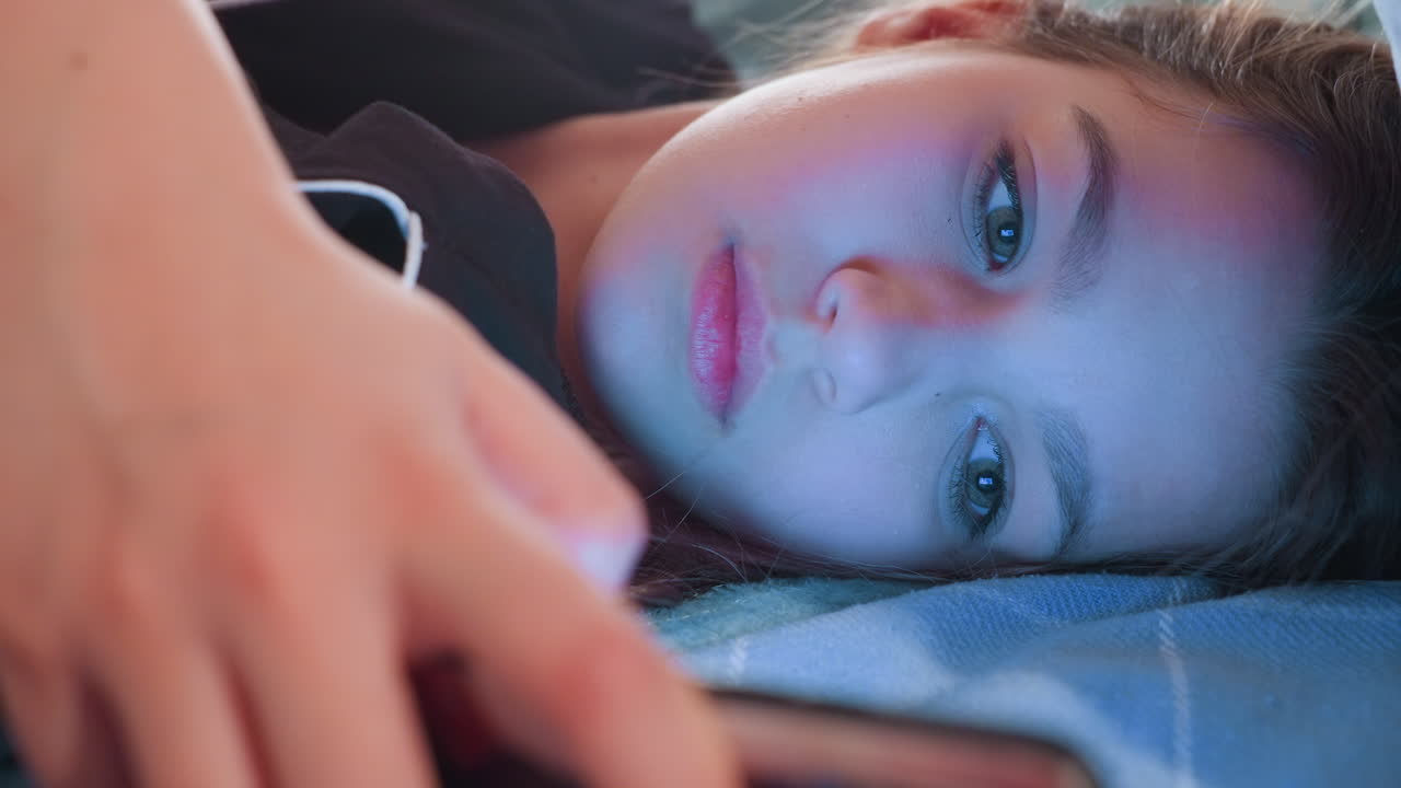 Young lady lying on bed while holding smartphone close to face and looking at screen with serious focused expression, scene shows digital interaction or leisure in cozy bedroom environment indoors
