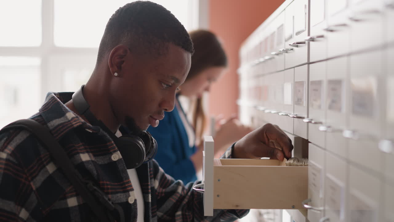 Visitors of library seek for library cards in paper storage. African American man with backpack wearing checkered shirt immersed in card index box. Students turn in books