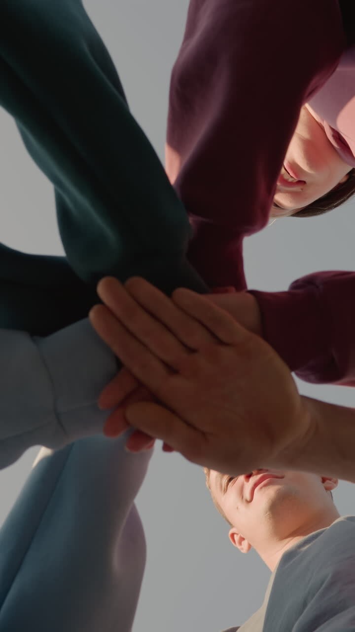 Upward view of people in hoodies placing their hands together as team members with sunlight creating a glowing effect around them, capturing team spirit and unity as they smile in outdoor setting