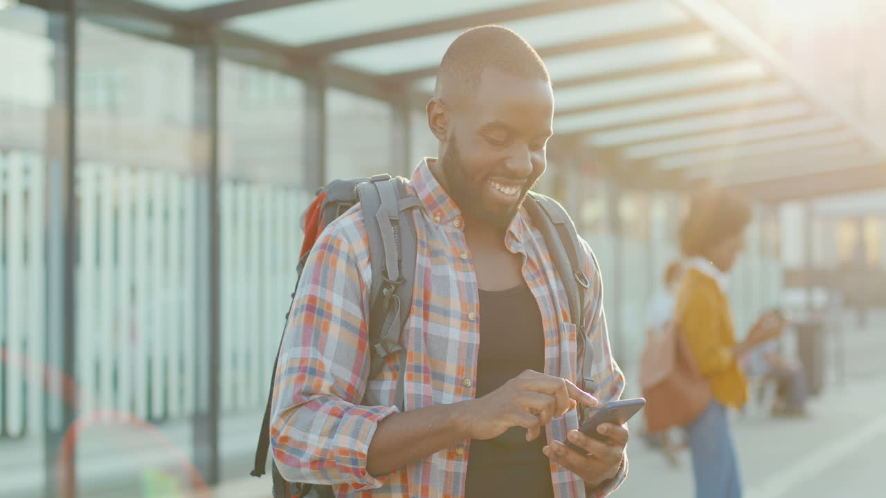 Young handsome African American man with backpack texting message on smartphone and smiling cheerfully at train station