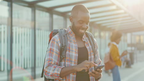 young handsome african american man with backpack texting message on smartphone and smiling cheerfully at train station