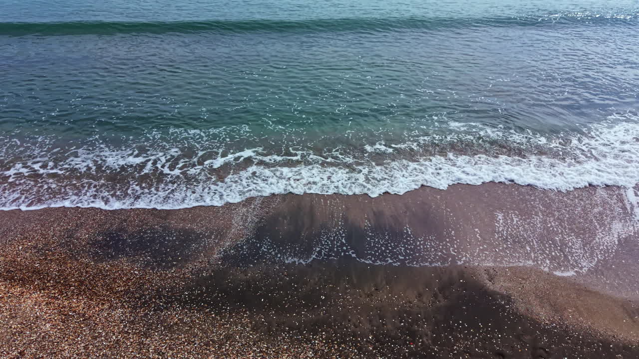 Onde che si infrangono sulla riva sabbiosa di una spiaggia nel pomeriggio, con acqua limpida e poche nuvole nel cielo