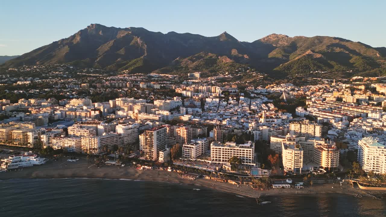 Vista aérea con dron a gran altitud de la ciudad costera de Marbella al atardecer, con el tranquilo mar Mediterráneo en primer plano y las montañas de Sierra Blanca al fondo, sobrevuelo costero suave