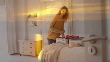 animation of happy caucasian woman packing suitcase in bedroom over sunset sky