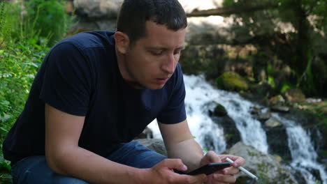 Joven-Solo-Navegando-Por-Las-Redes-Sociales-En-Un-Teléfono-Y-Fumando-Un-Cigarrillo-Cerca-De-Una-Cascada