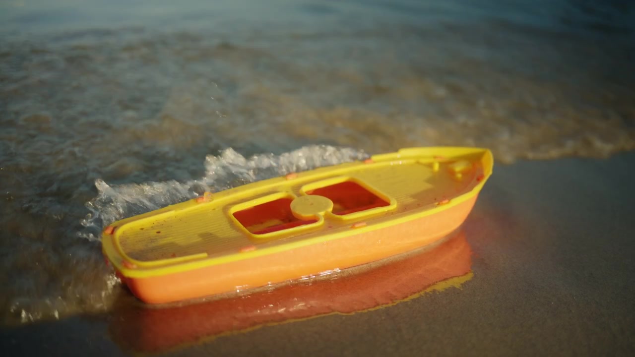 Una barchetta giocattolo di plastica gialla e arancione che riposa sulla sabbia bagnata in riva all'acqua, con piccole onde che si infrangono dolcemente.