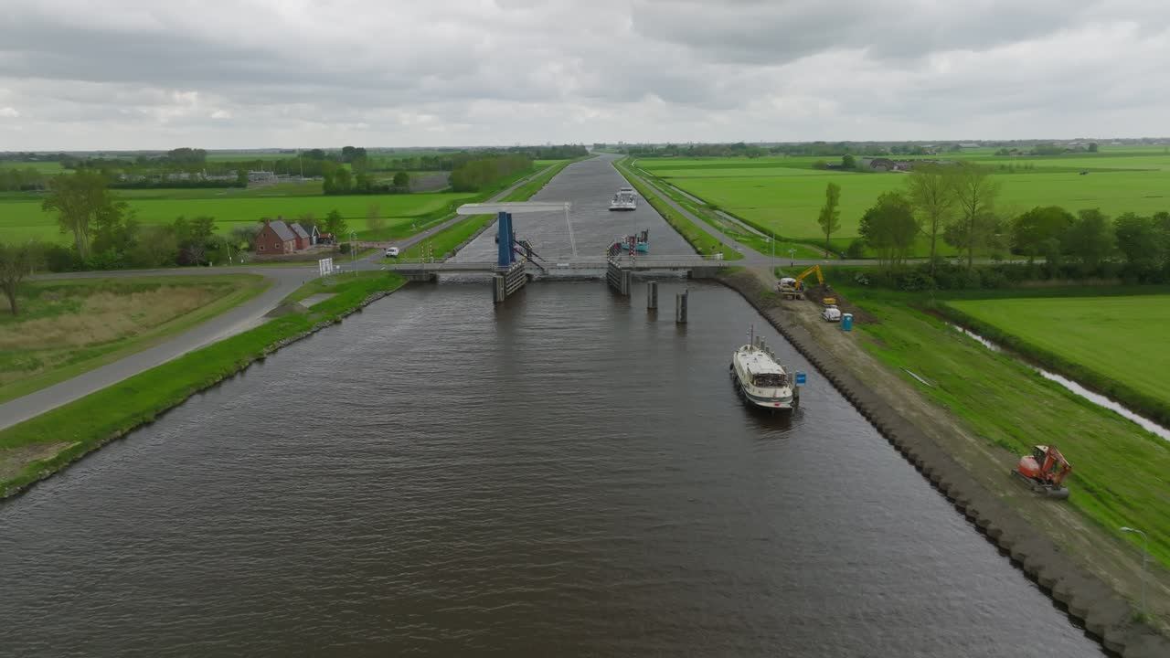 Approach view toward a bascule (draw) bridge on a straight Dutch canal, with farmland, service road and small craft visible under grey skies