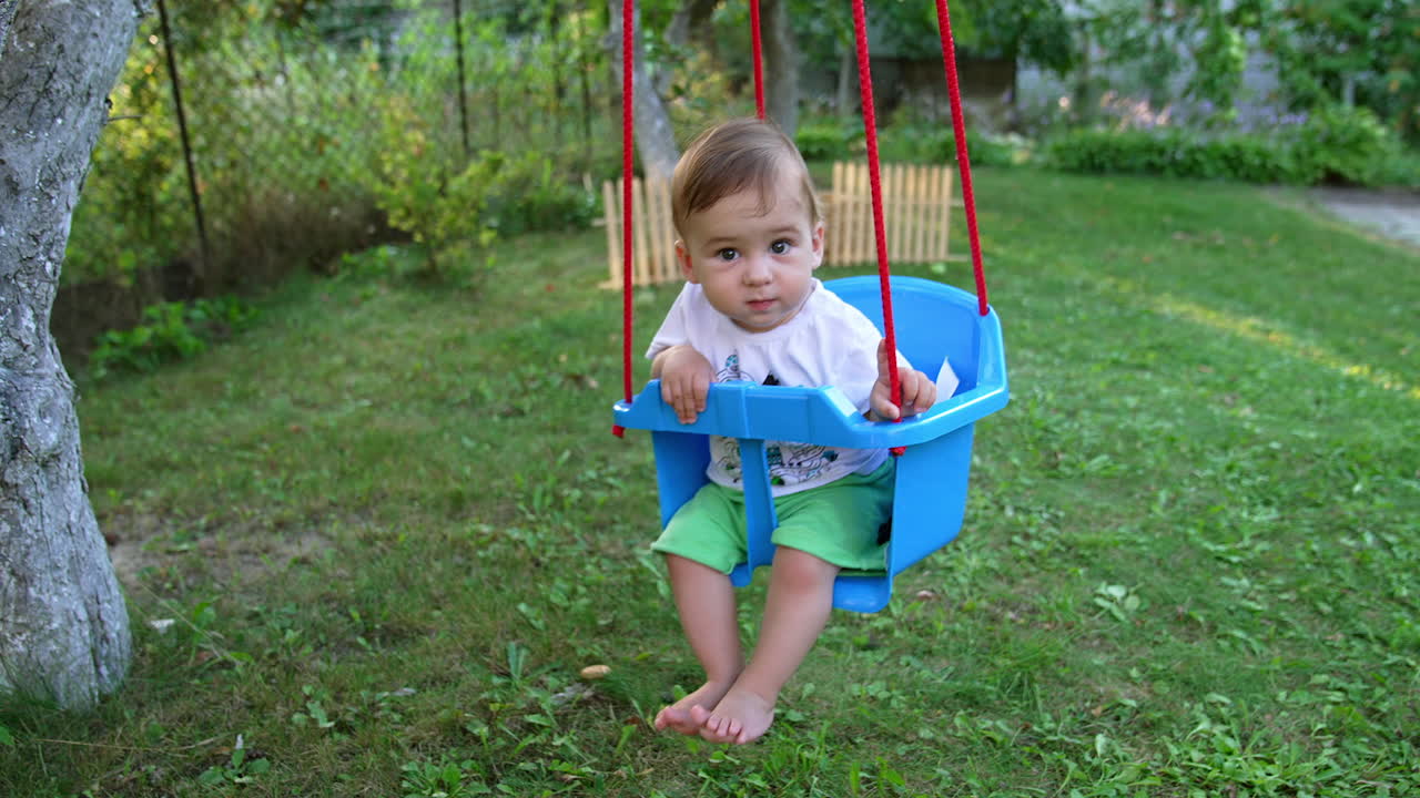 Lovely calm baby boy swinging in a swing looking into camera with his eyes wide open. Adorable barefoot Caucasian kid swaying in the garden in summer.