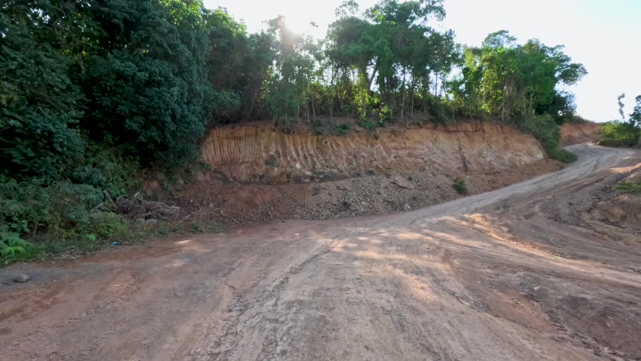 Vehicle travels along winding dirt road through tropical hillside, sun rays filtering through dense trees