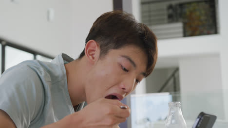 asian male teenager sitting at table with smartphone and having breakfast