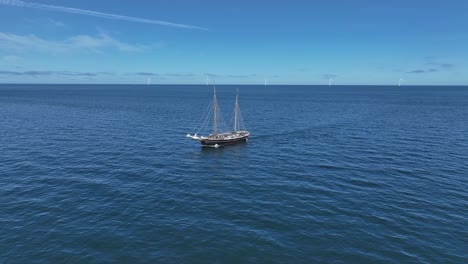 tall ship sailing through calm blue sea with blue sky and wind turbines on the horizon