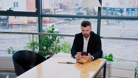 bearded man in black suit sitting at the table. businessman using phone searching for information and shows the result of search.