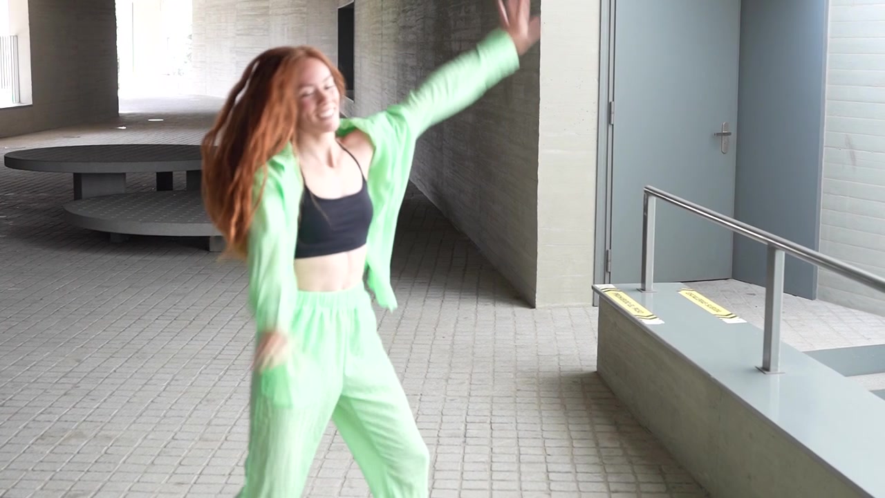 En glad kvinde danser energisk i et iøjnefaldende grønt outfit ved siden af en bytrappe