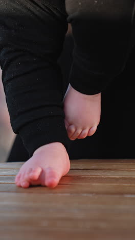 tiny bare feet of a baby on the wooden surface. kid wearing black pants stepping by the table. blurred backdrop. vertical video