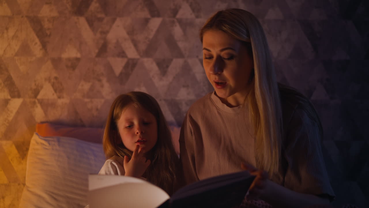 una niña escucha a su madre leyendo un libro en un dormitorio oscuro