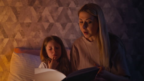 una niña escucha a su madre leyendo un libro en un dormitorio oscuro