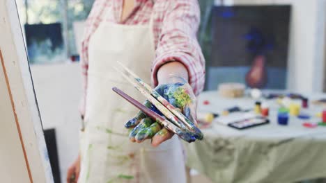 hand of biracial female artist in apron holding brushes in painting studio, slow motion