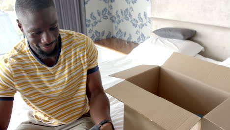 unpacking clothes from cardboard box, man smiling and sitting on bed