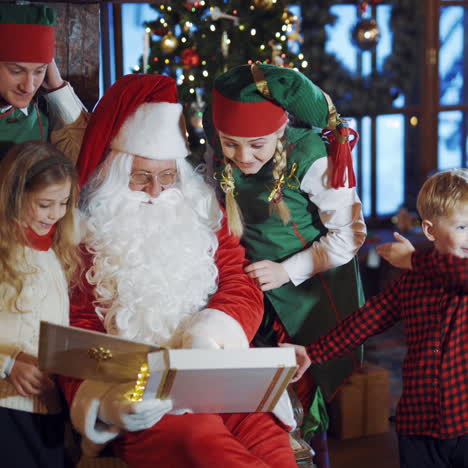 fairy characters and children at christmas indoors. santa claus is sitting with album and kids, funny elves standing nearby in the room with christmas decorations.