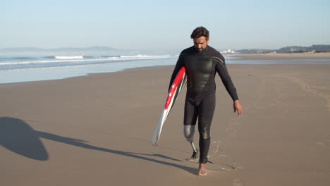 long shot of a male surfer with artificial leg walking along beach and holding surfboard under arm 1