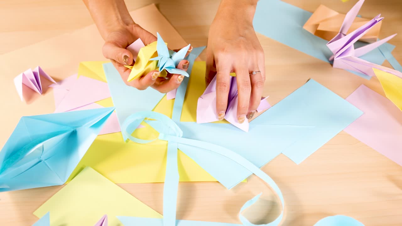 Person folds pastel origami cranes on wooden table, surrounded by paper, under bright lighting