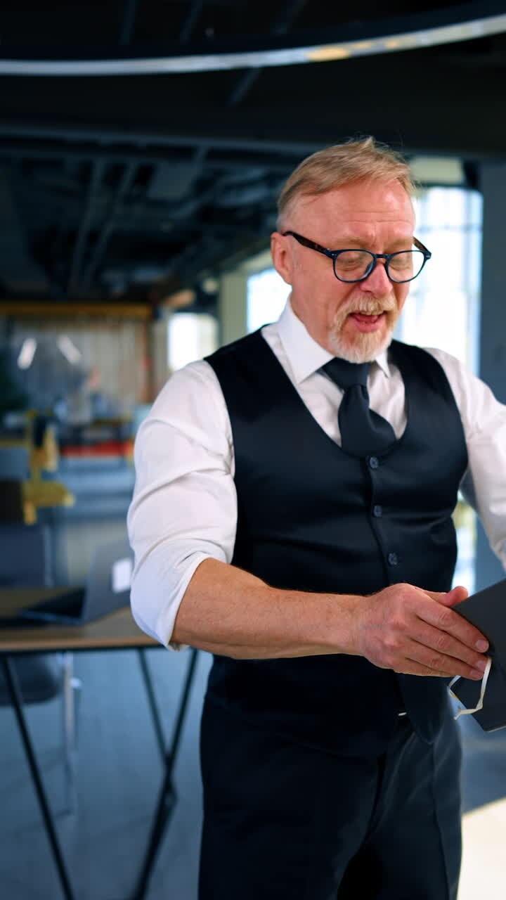 Caucasian old man wearing white shirt, trousers, vest and tie stands in the office. Man puts a notebook on his head and stands smiling. Vertical video.