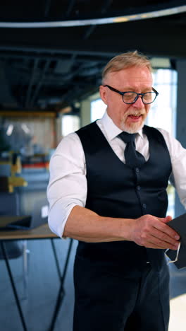 caucasian old man wearing white shirt, trousers, vest and tie stands in the office. man puts a notebook on his head and stands smiling. vertical video.