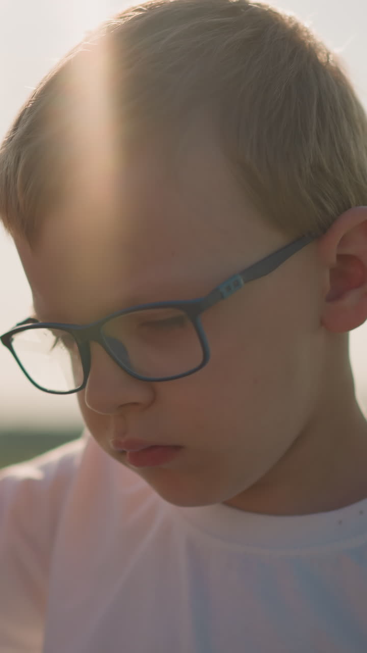 A young boy wearing a white top and glasses, with a red band on his wrist, is sitting in a grassy field. He gently touches his head and appears sleepy, bathed in soft sunlight