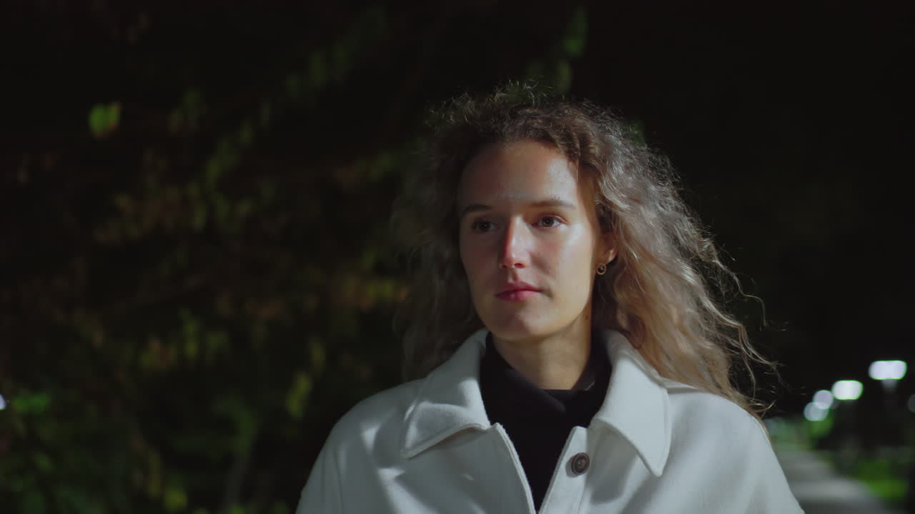 Serious woman with curly hair in white coat walks outdoors at night, illuminated by soft lighting against dark background with blurred greenery, reflecting thoughtful expression during evening stroll