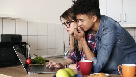 una chica de aspecto caucásico y su novio mulato, ambos en sus 20 años, usando una computadora portátil, navegando por la web en la cocina de su casa. él la abraza tiernamente por la espalda.