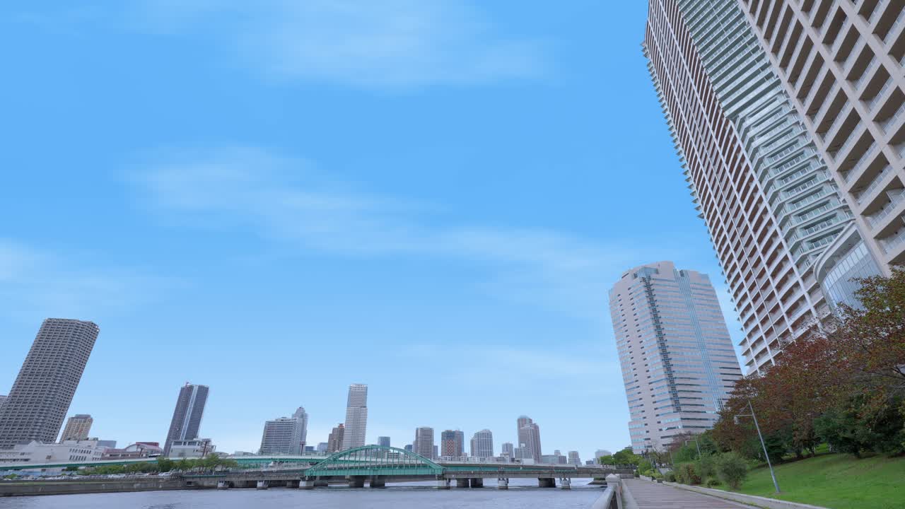 A dramatic low-angle shot of the Tokyo skyline from a riverbank, with tall skyscrapers and a clear blue sky
