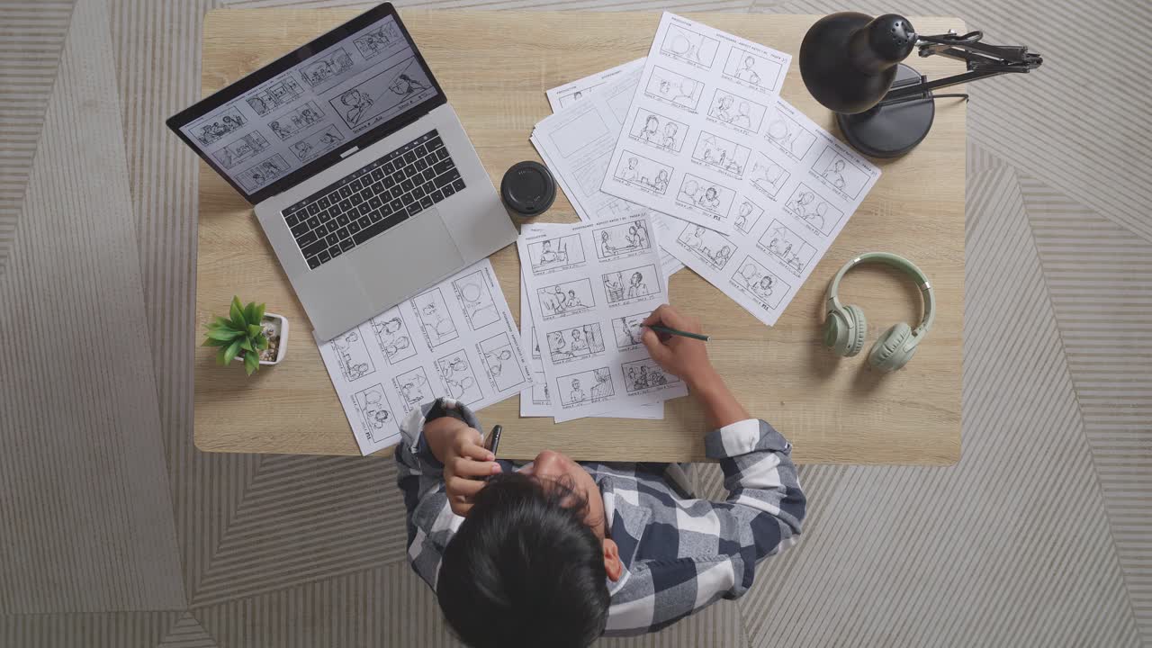 vista superior de un artista masculino asiático hablando en un teléfono inteligente mientras dibuja un guión gráfico para la película en la mesa con una computadora portátil y auriculares en el estudio