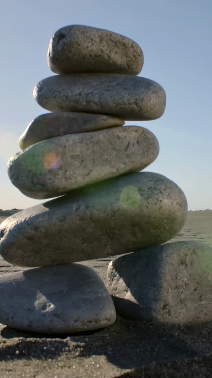 Vertical video: Hand placing pebbles on beach, balancing stone tower with lens flare for design