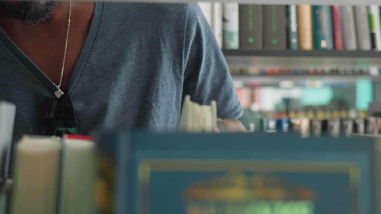 Close up of adult in grey t-shirt and necklace standing near bookshelf in library, partially obscured by blurred row of books in foreground, appearing to search for specific title among collection