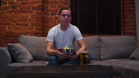 the young man is sitting on the couch, watching tv and eating chips in his living room