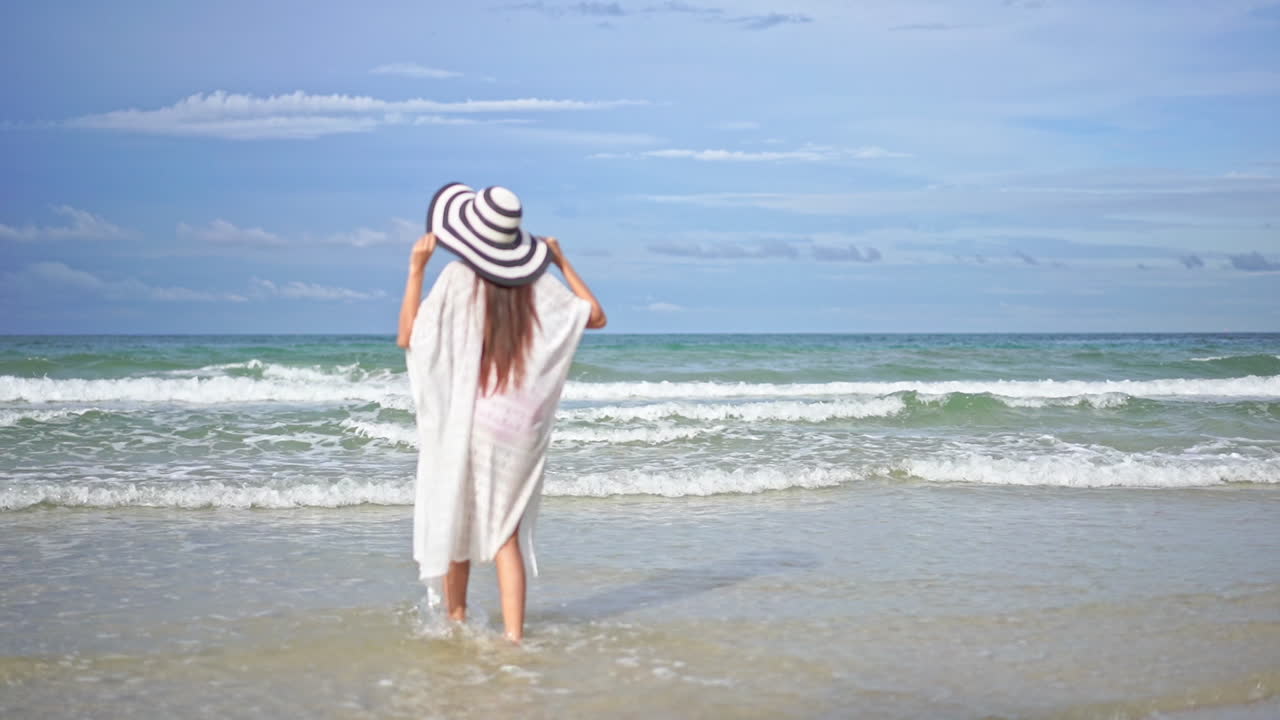 하얀 여름 튜닉을 입은 흥분한 여성의 뒷모습과 카리브해 모래사장에서 파도를 향해 달려가는 모자, 느린 동작