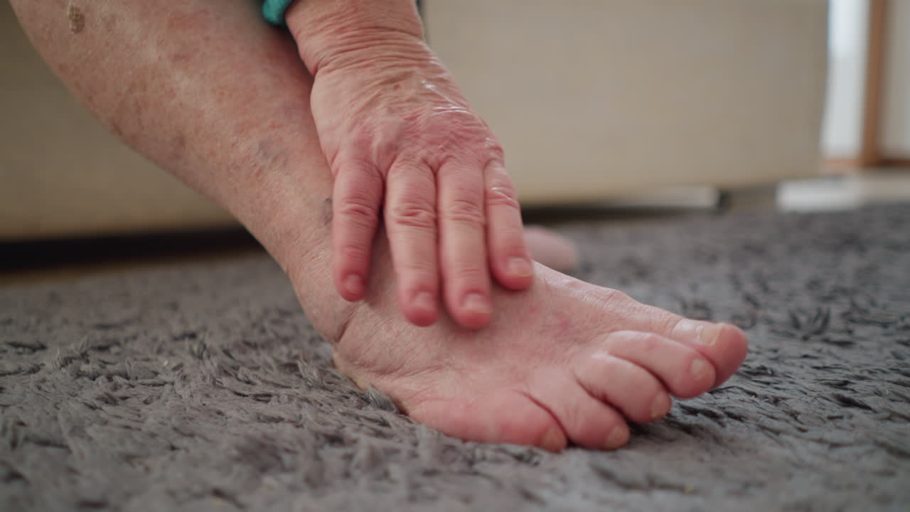 old woman in green sitting on bed, applying ointment to leg. She is massaging it with one hand while holding the tube with the other. Soft lighting highlights her effort in self-care