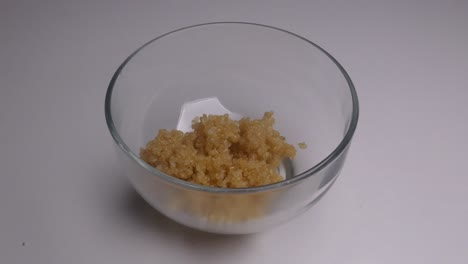 a clear glass bowl in a white background. a hand continuously pours in more and more cooked white peruvian quinoa into the bowl using a stainless steel spoon