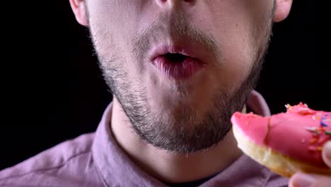 close-up portrait of man biting and chewing delicious glazed pink donut with colorful chips and eyes on black background.